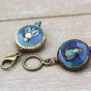 May include: Two round, blue and gold, bee-themed charms with a bronze clasp and ring. The charms are made of glass and have a detailed bee design.