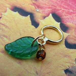 May include: A gold charm with a green glass leaf and a brown glass bead. The charm is attached to a gold ring.