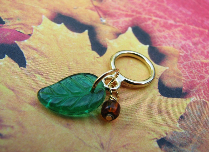 May include: A gold charm with a green glass leaf and a brown glass bead. The charm is attached to a gold ring.