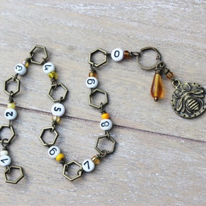 May include: A handmade bronze-toned keychain with a bee charm, a teardrop bead, and a chain of hexagonal links. The chain has white beads with black numbers from 0 to 9. The keychain is on a wooden surface.