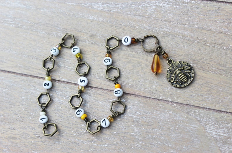 May include: A handmade bronze-toned keychain with a bee charm, a teardrop bead, and a chain of hexagonal links. The chain has white beads with black numbers from 0 to 9. The keychain is on a wooden surface.