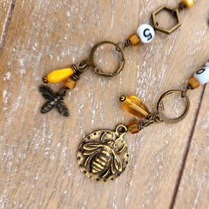 May include: Close-up of a handmade bee-themed jewelry piece. The piece features a bronze-colored bee charm, yellow and amber-colored beads, and a small metal bee. The jewelry is on a wooden surface.