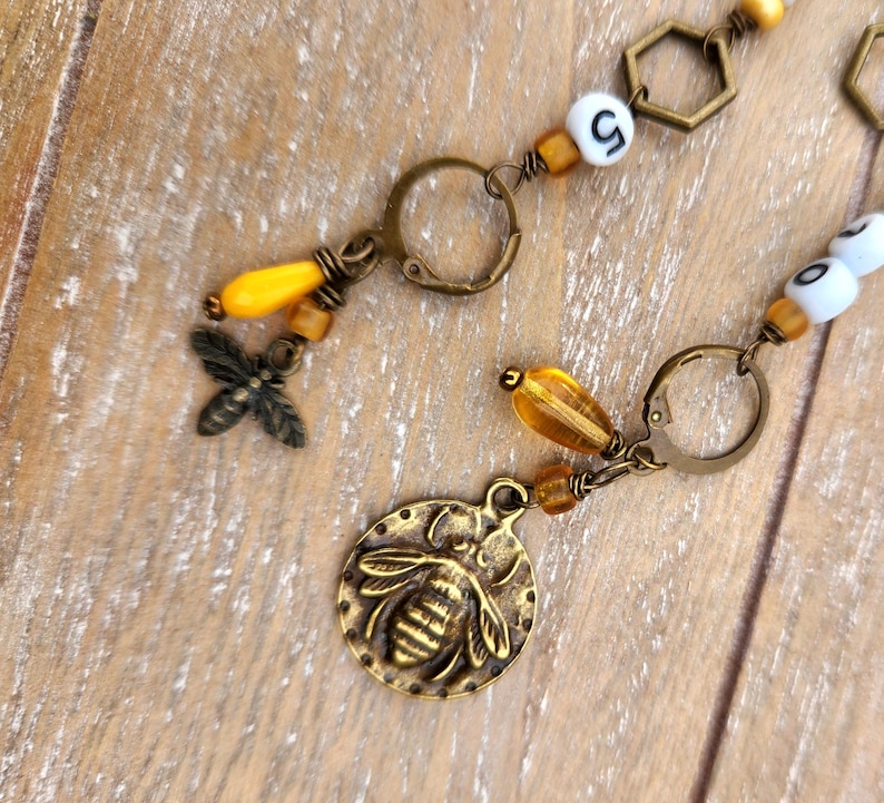 May include: Close-up of a handmade bee-themed jewelry piece. The piece features a bronze-colored bee charm, yellow and amber-colored beads, and a small metal bee. The jewelry is on a wooden surface.