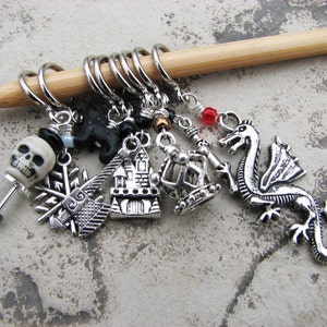 May include: A set of eight silver knitting stitch markers with various charms, including a skull, a snowflake, a house, a crown, a dragon, and a black cat. The markers are attached to a wooden knitting needle.