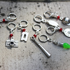 May include: A set of ten silver-toned stitch markers with red beads and various charms, including a bicycle, a camera, a number eleven, a baseball bat, a compass, a lightbulb, and a hat.