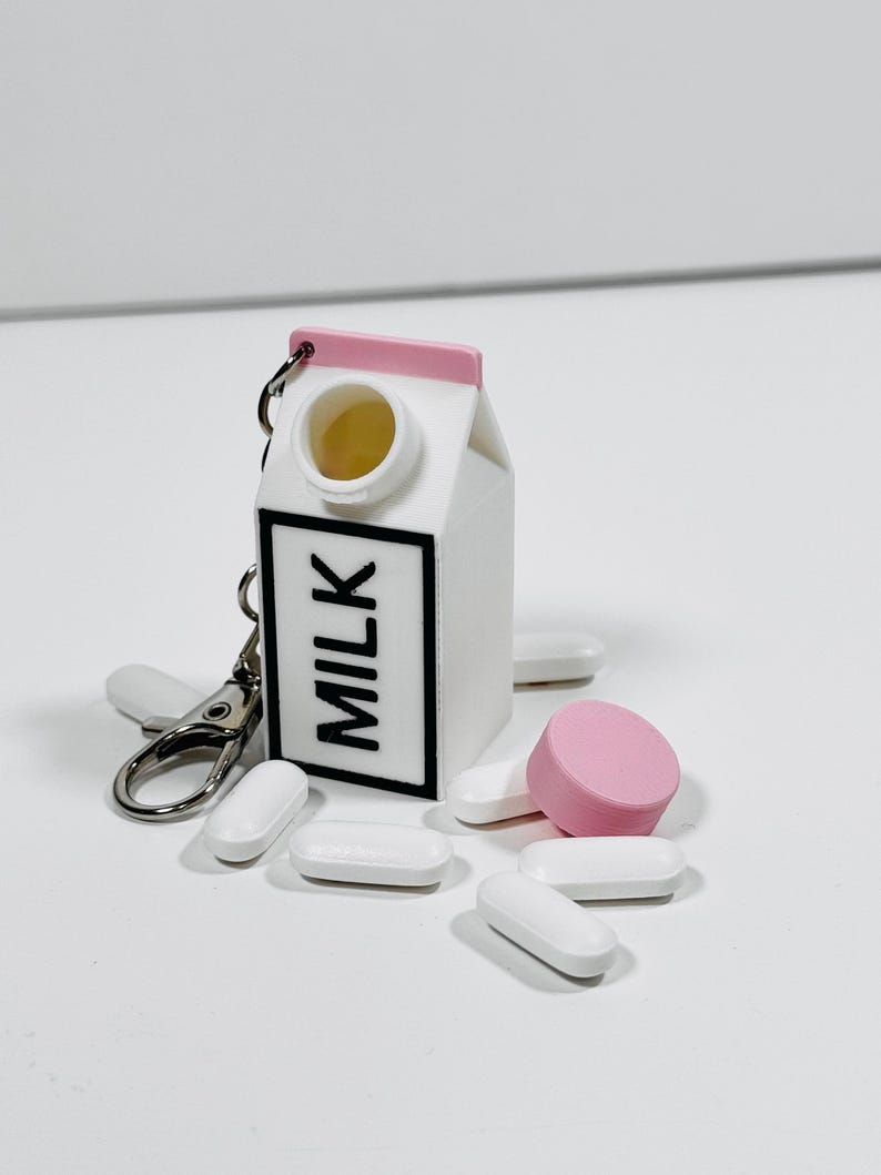 Op de afbeelding: Een witte melkpak sleutelhanger met een roze deksel en een zwart "MILK" label. De sleutelhanger is open en er liggen verschillende witte pillen en &eacute;&eacute;n roze pil rondom.