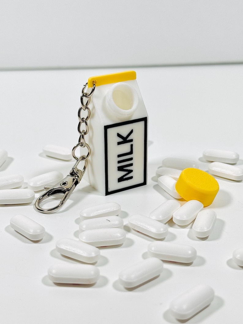 Op de afbeelding: Een witte melkpak sleutelhanger met een geel deksel en een zilveren ketting. De sleutelhanger is open en er liggen witte pillen verspreid eromheen. Het deksel van de sleutelhanger is geel en ligt naast de sleutelhanger.