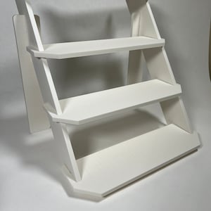 Puede incluir: Estantería blanca de tres niveles con un diseño inclinado. La estantería está hecha de madera y tiene un acabado blanco.