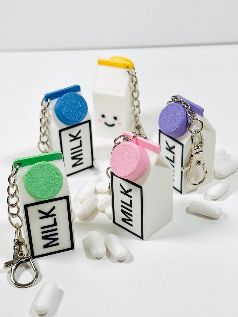Op de afbeelding: Vier miniatuur melkpak sleutelhangers met verschillende gekleurde deksels: groen, blauw, roze en paars. Elke sleutelhanger heeft een zilveren ketting en een zilveren ring. De pakken zijn wit met zwarte letters die "MELK" zeggen.