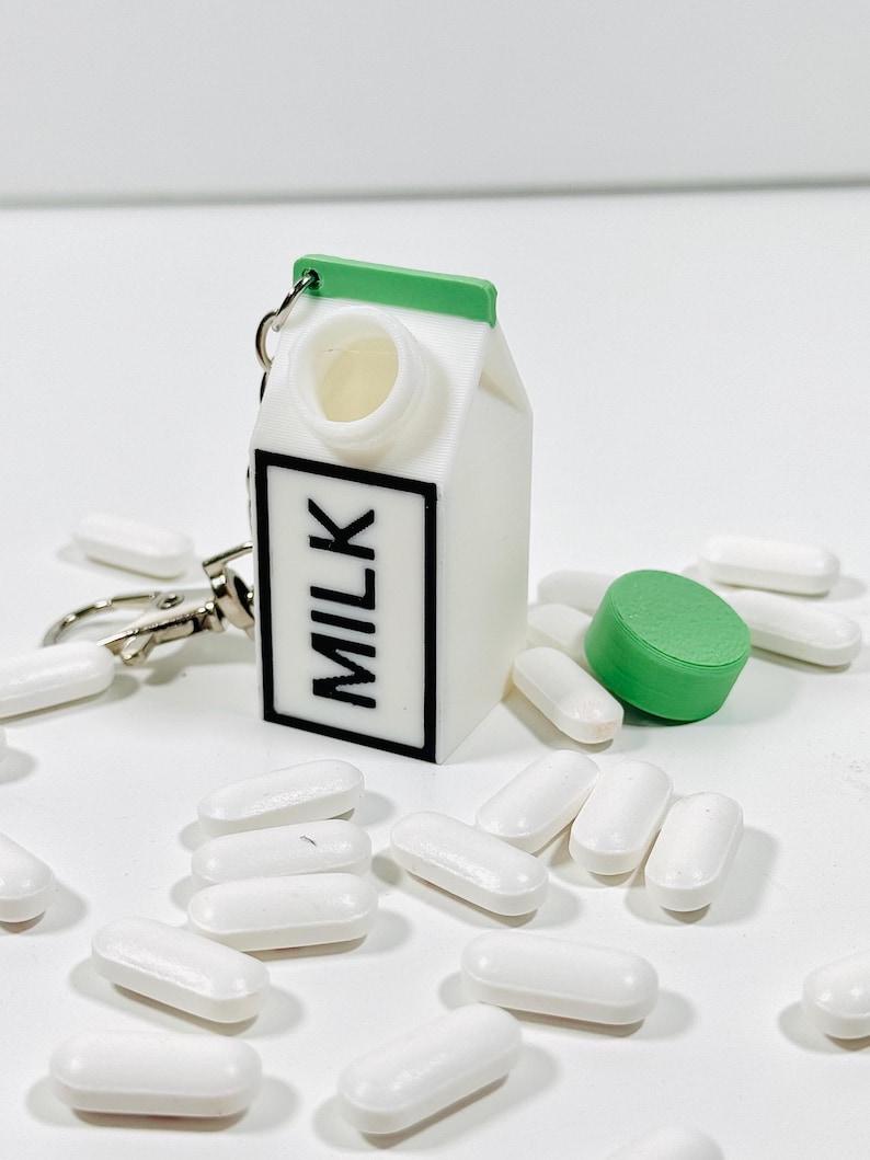 Op de afbeelding: Witte melkpak sleutelhanger met een groen deksel en een zwart "MILK" label. De sleutelhanger is omringd door witte pillen.