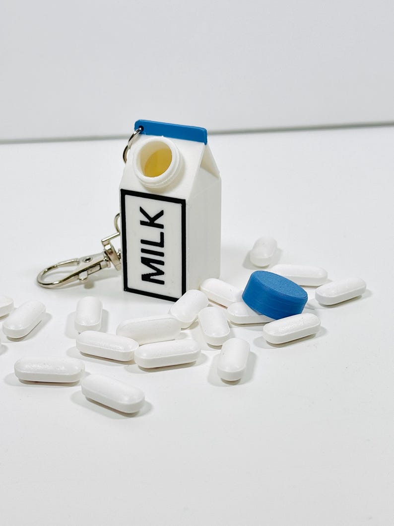 Op de afbeelding: Een witte melkpak sleutelhanger met een blauw deksel en het woord "MILK" gedrukt op de voorkant. De sleutelhanger is omringd door witte pillen.