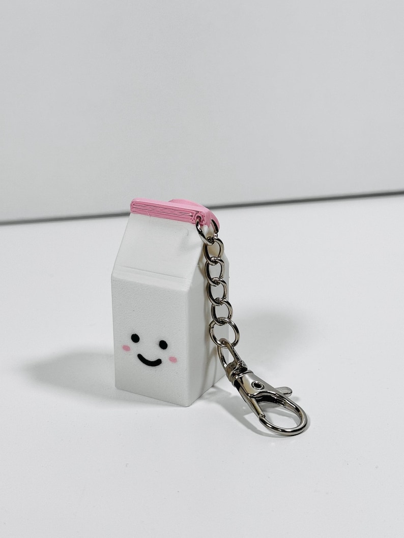 Op de afbeelding: Een witte melkpak sleutelhanger met een roze deksel en een glimlachend gezicht. De sleutelhanger heeft een zilveren metalen ketting en een zilveren sluiting.