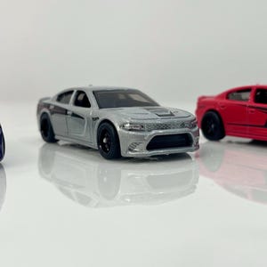Peut inclure: Trois voitures miniatures alignées. La première est violet foncé, la seconde argentée et la troisième rouge. Les trois voitures ont des roues noires et sont sur une surface blanche.