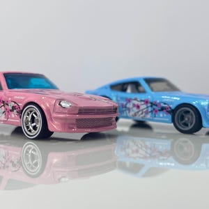 May include: Two toy cars, a pink and a blue, are displayed. The pink car has a cherry blossom design and white-walled tires. The blue car also features a cherry blossom design. Both cars have a sleek, sporty design.