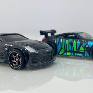 May include: Two black toy cars with spoilers. One car has a detailed design on the side, while the other has a blue and green geometric pattern. Both cars have black wheels and are on a white surface.