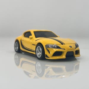 Puede incluir: Un coche de juguete amarillo con rayas de carreras negras y la palabra "Gazoo" en el lateral. El coche es un modelo de un Toyota Supra.