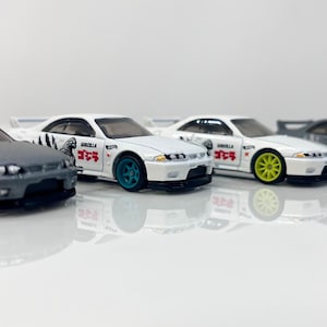 May include: Four toy cars in a row, each with a different color scheme. Two are gray with gold wheels, and two are white with colored wheels. Each car has a Godzilla graphic and Japanese text.