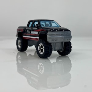 Puede incluir: Una camioneta de juguete negra y roja con neumáticos todoterreno negros grandes. La camioneta tiene un parachoques delantero gris y un parabrisas azul. El lateral de la camioneta dice "Parts Delivery" en texto blanco con rayas rojas y blancas.