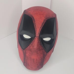 May include: A red and black Deadpool mask with a textured surface. The mask has a black eye area with white eyes.