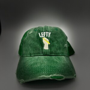 May include: A green distressed baseball cap with a white embroidered logo that says "Lefty" and a yellow pencil in a fist.
