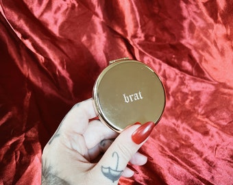 hand engraved gold compact mirror with the word “brat” in an old English style lettering.