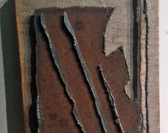 May include: A small, rectangular piece of art with a brown wooden frame. The artwork is made of metal and has a rusty brown color. There are three thin, silver lines running diagonally across the metal. The metal is slightly raised from the frame.