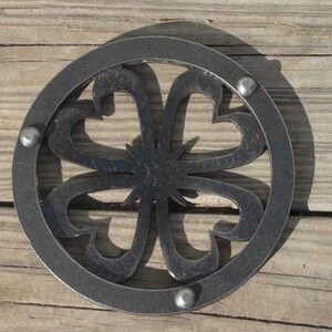 Könnte beinhalten: Ein kreisförmiger, dunkelgrauer Metall-Topfuntersetzer mit dekorativem Blumendesign. Der Untersetzer hat ein zentrales Sternmuster, umgeben von stilisierten, gebogenen Formen. Drei Nieten sichern den äußeren Ring. Zum Schutz von Oberflächen vor heißem Kochgeschirr.