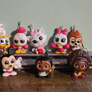 May include: A collection of eight character figurines, including Mickey Mouse, Donald Duck, Daisy Duck, Chip, and Marie. Each figure is brightly colored and has a small loop for hanging. The figurines are displayed on a gray shelf.