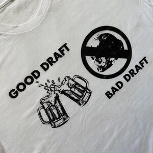 May include: White t-shirt with a black graphic design. The design includes two beer mugs clinking, the words "GOOD DRAFT" and "BAD DRAFT", and a circular image of a skull with a line through it. The shirt is made of a soft material.