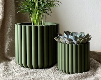 May include: Two green ceramic planters with a ribbed design. The larger planter holds a green leafy plant, while the smaller planter holds a succulent.