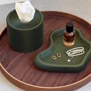 Op de afbeelding: Een groene dienblad met een geribbelde textuur, een klein flesje parfum, een parelketting en twee gouden oorringen. Het dienblad ligt op een houten oppervlak.