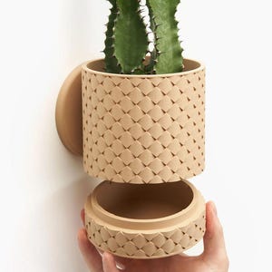 Op de afbeelding: Een beige keramische plantenpot met een geometrisch patroon, met een afneembaar deksel. De pot is aan een muur bevestigd en bevat een groene cactusplant.