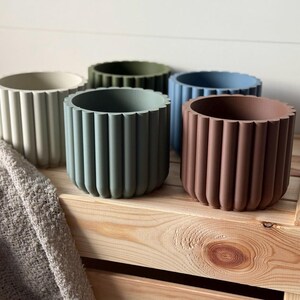 May include: Five different colored concrete planters with a ribbed design. The planters are arranged on a wooden shelf.