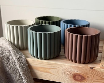 Könnte beinhalten: Fünf unterschiedlich farbige Beton-Blumentöpfe mit geripptem Design. Die Töpfe sind in einer Reihe auf einem Holzregal angeordnet. Die Farben sind beige, grün, blau, braun und grün.