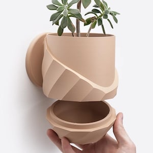 May include: A beige ceramic planter with a geometric design. The planter is split into two sections, with the top section holding a plant and the bottom section being a separate container. The planter is mounted to a wall.