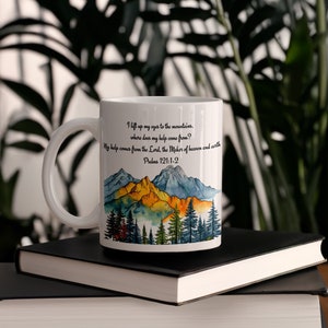 Op de afbeelding: Een witte keramische mok met een aquarel-illustratie van bergen en bomen. De mok heeft een zwart handvat en de tekst "I lift up my eyes to the mountains, where does my help come from? My help comes from the Lord the Maker of heaven and earth Psalms 121:1-2" gedrukt op de zijkant.