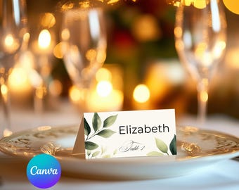 Pode incluir: Um cartão de lugar branco com folhagem verde e o nome "Elizabeth" e "Mesa 1" escritos em caligrafia preta.