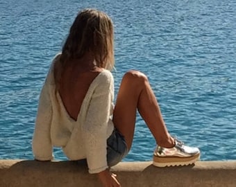 Könnte beinhalten: Eine Frau in einem weißen Strickpullover mit offenem Rücken sitzt auf einer Betonmauer und blickt auf eine blaue Wasserfläche. Sie trägt Jeans-Shorts und silberne Plateauschuhe.