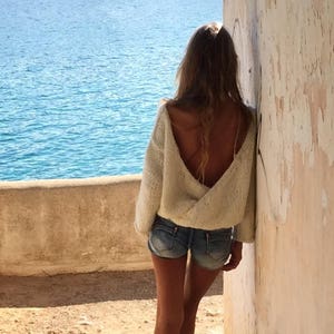 May include: A woman wearing a white knitted sweater with an open back and denim shorts stands near a wall overlooking a body of water.