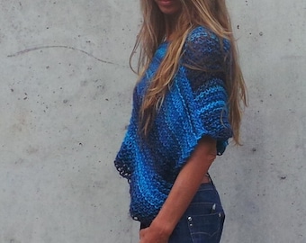 Puede incluir: Una mujer con un poncho de punto azul y negro con pantalones cortos de mezclilla cortados. Está de pie frente a una pared de concreto.