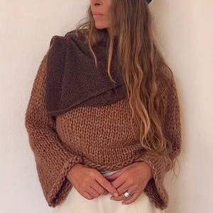 Puede incluir: Un suéter de punto marrón con una bufanda a juego y una gorra azul oscuro. El suéter tiene mangas anchas y se combina con pantalones color crema. El conjunto es de estilo casual y acogedor.