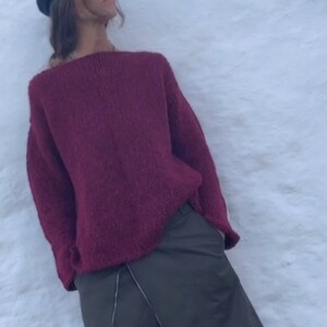 May include: A woman wearing a burgundy knitted jumper and a dark green skirt with a zip detail. She is wearing a black newsboy cap and black loafers.