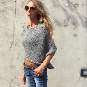 May include: A grey knitted sweater with a loose fit and short sleeves. The sweater is cropped and has a slightly uneven hemline.