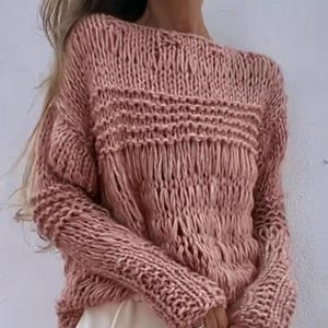 Puede incluir: Un suéter de punto grueso de color rosa polvoriento con detalles de flecos. El suéter presenta un escote amplio y mangas largas. El patrón de punto incluye bandas horizontales y secciones de tejido abierto, creando un aspecto texturizado. La prenda está hecha de un material suave y cómodo.