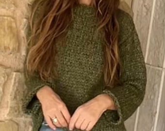 May include: A woman wearing a green knit sweater and blue jeans. The sweater has long sleeves and a loose fit. The woman is leaning against a stone wall.