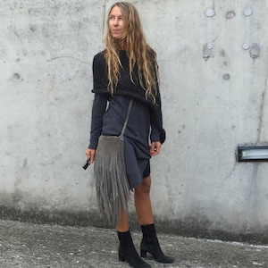 May include: A woman wearing a grey knitted poncho and a grey dress with a fringe bag slung over her shoulder. She is wearing black ankle boots.