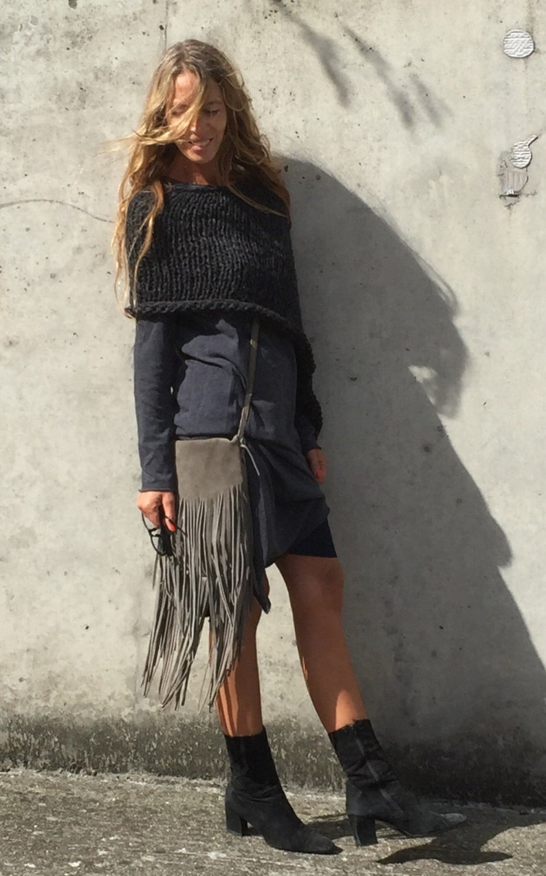 May include: A woman wearing a dark grey knitted poncho over a dark blue dress. She is carrying a grey suede fringe bag with a long strap. She is wearing black ankle boots.