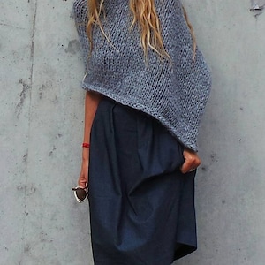 May include: A grey knitted poncho with a loose, flowing fit. The poncho is worn over a dark blue skirt.