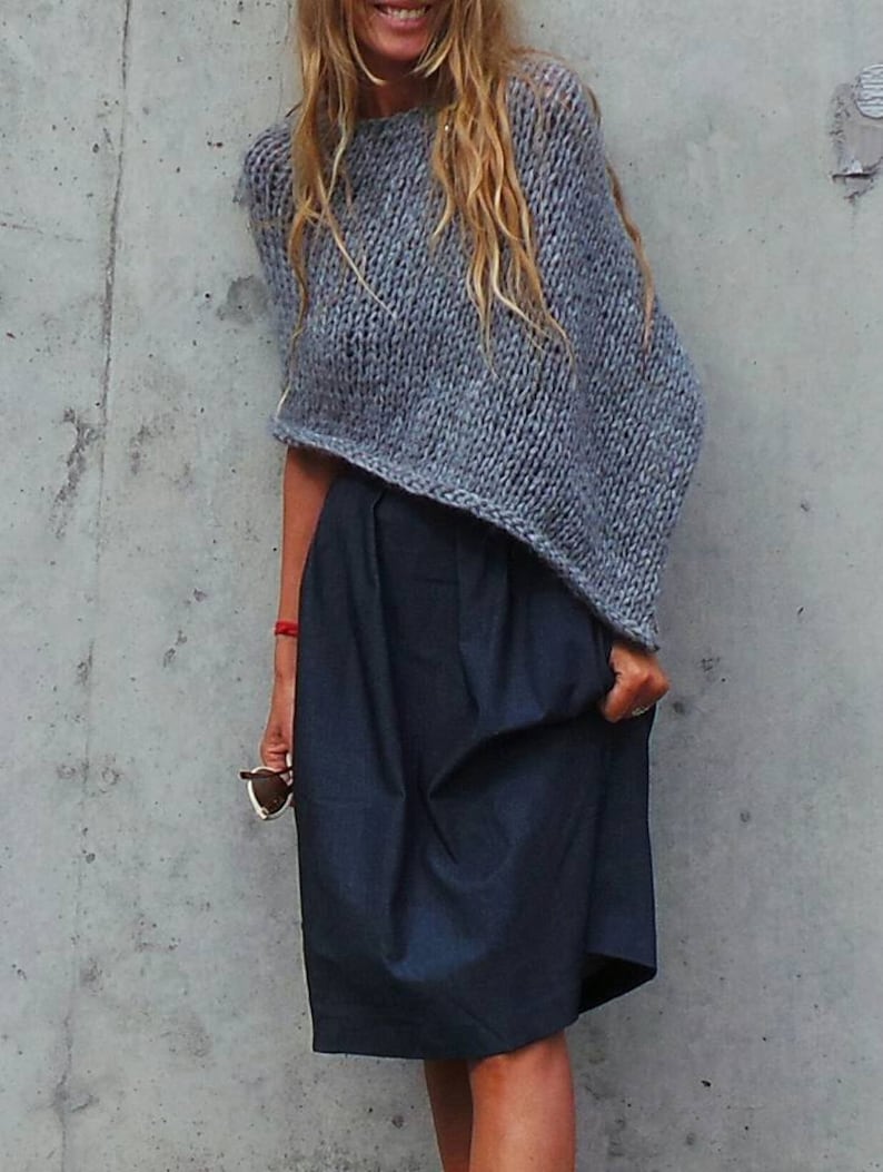 May include: A grey knitted poncho with a loose, flowing fit. The poncho is worn over a dark blue skirt.