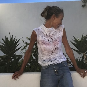 Könnte beinhalten: Eine Frau trägt ein weißes Häkeltop und eine blaue Jeans. Sie steht vor einer weißen Wand mit Pflanzen auf beiden Seiten.